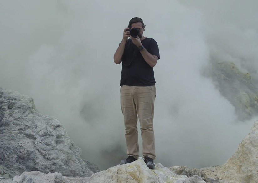 A man stands on rocks with stream rising behind him. He is taking a photo faced toward the viewer.