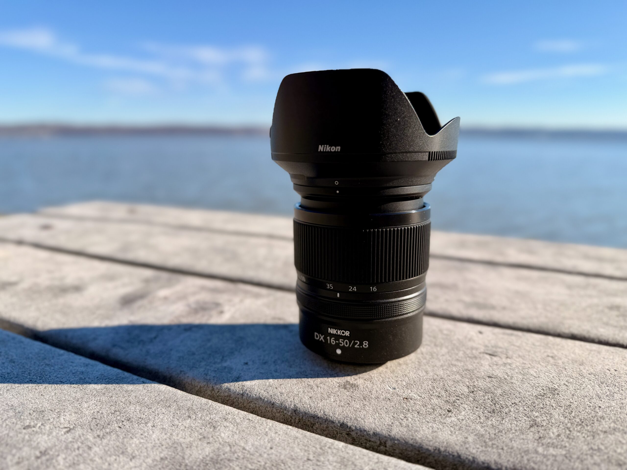 The Nikon 16-50mm f/2.8 VR on a park bench with blue skies and a reiver behind it. 
