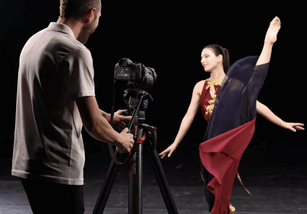 A ballerina being photographed against a black bacground by a photographer with camera on a tripod